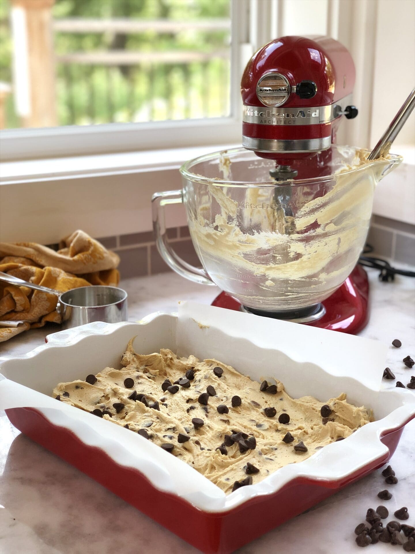 A cherry red stand mixer streaked with batter sits by a cottage window.  A pan of chocolate chip blondies sits in the foreground, ready for the oven.