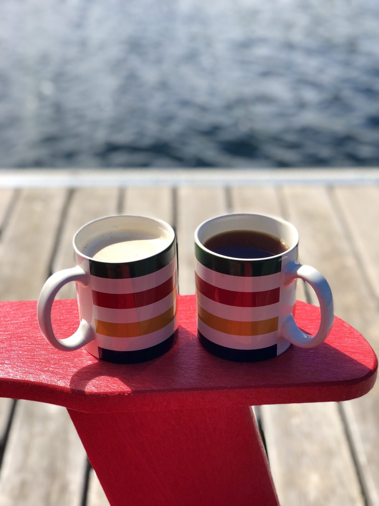 A cup of tea and a cup of coffee sit together on the arm of a red muskoka chair.  The chair sits on a dock, with the blue water of the lake visible in the background.