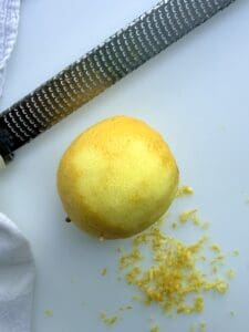 A fresh lemon is seen from above with a zester and a pile of fresh lemon zest.