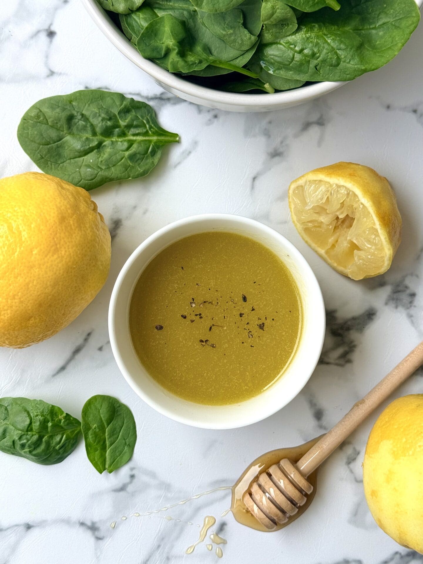 A bowl of Mediterranean Lemon Vinaigrette is seen from above, surrounded by fresh lemons, honey and baby spinach.