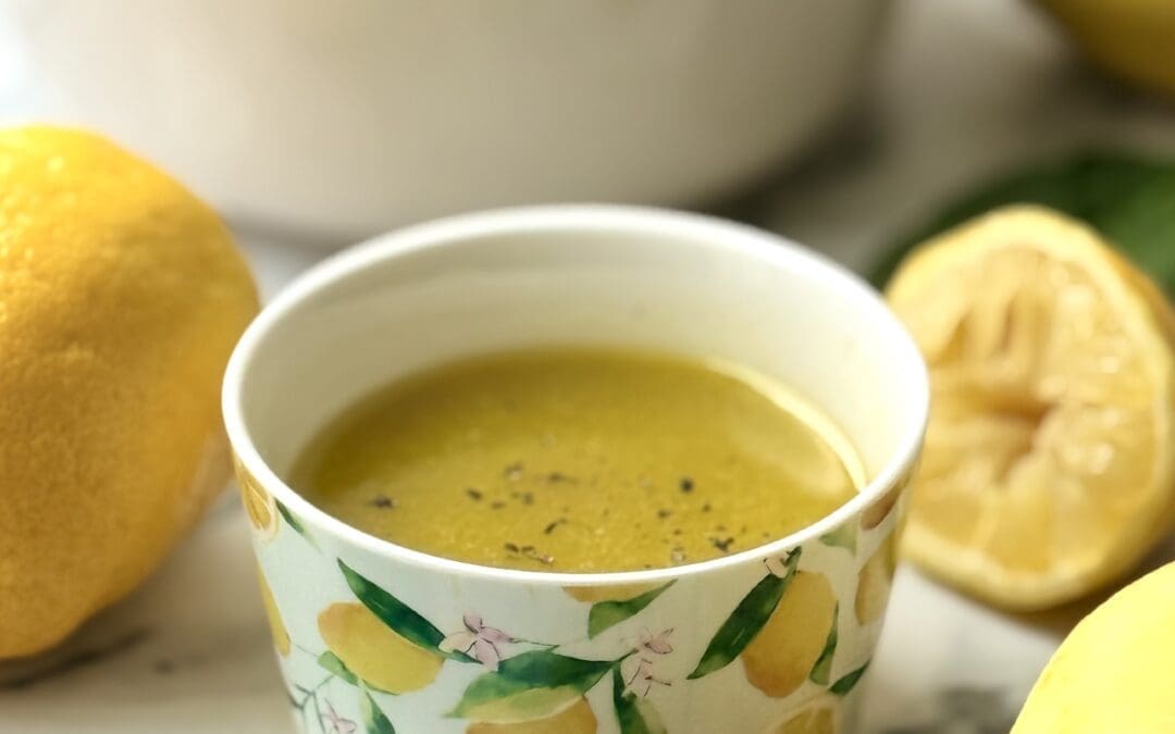 A decorative bowl with a vibrant lemon print is filled with Mediterranean Lemon Vinaigrette. The bowl of dressing is surrounded by fresh lemons, honey and greens.