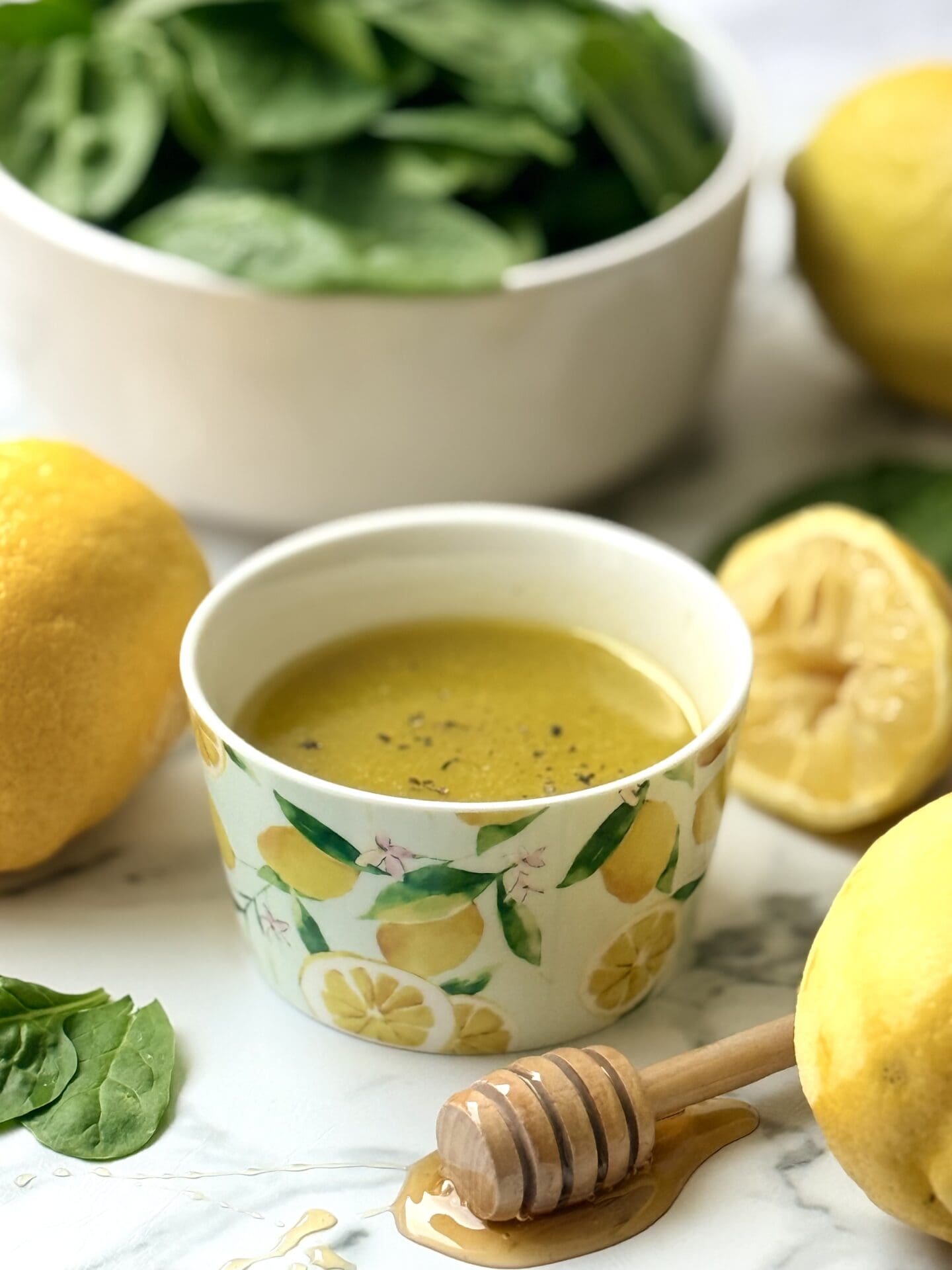 A decorative bowl with a vibrant lemon print is filled with Mediterranean Lemon Vinaigrette. The bowl of dressing is surrounded by fresh lemons, honey and greens.