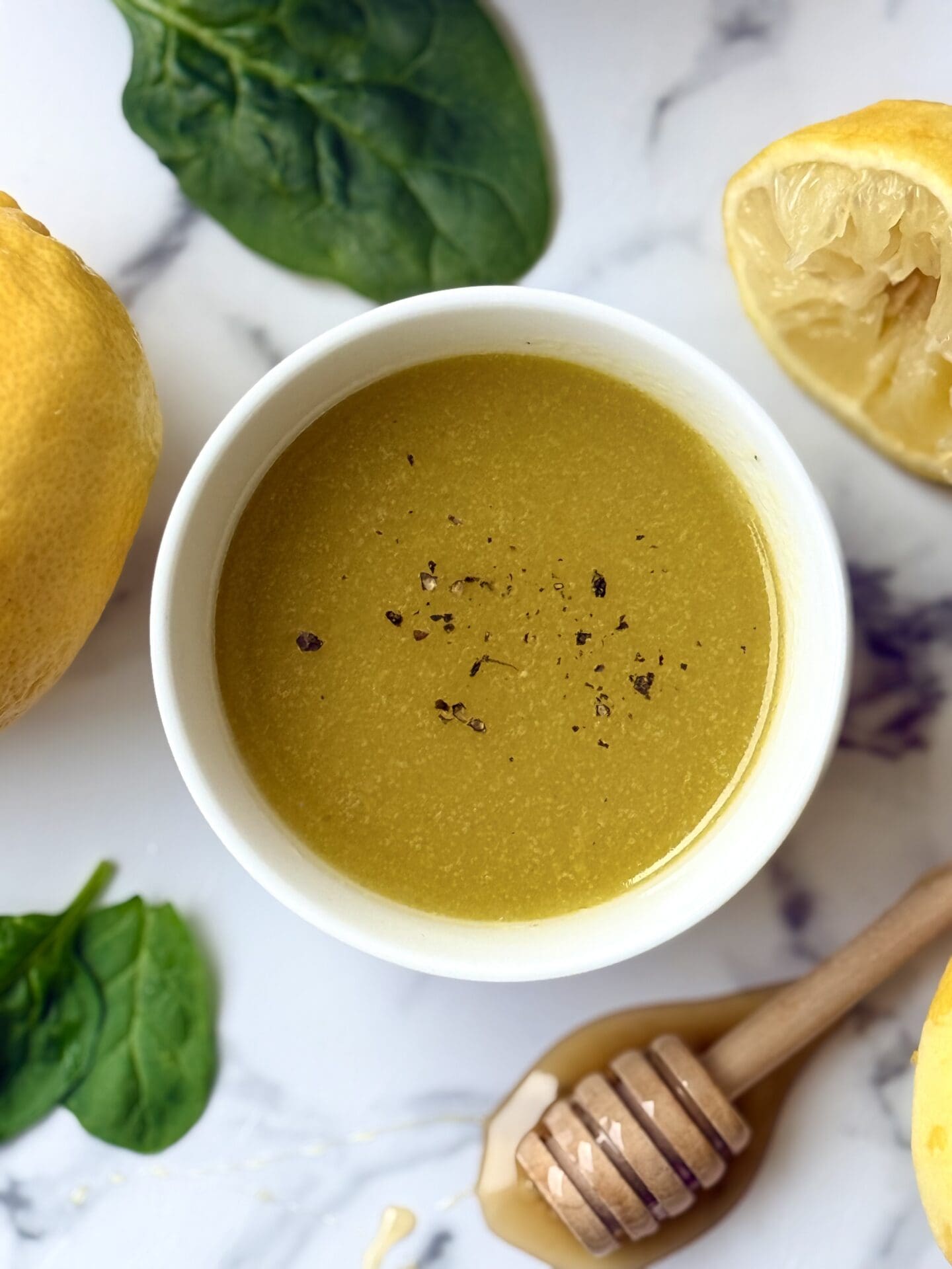 A bowl of Mediterranean Lemon Vinaigrette is seen from above on a white marble counter. Fresh lemons, a honey dipper and baby spinach leaves surround the bowl.