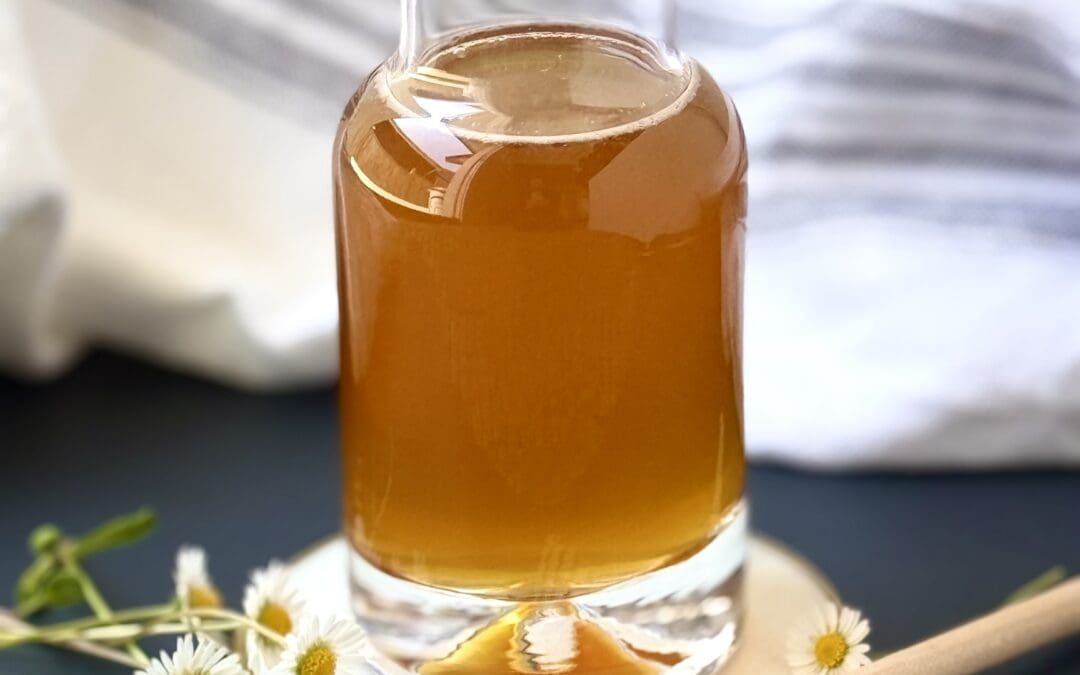 A glass bottle of honey simple syrup sits on a white and gold coaster, surrounded by tiny daisies and a honey dipper.
