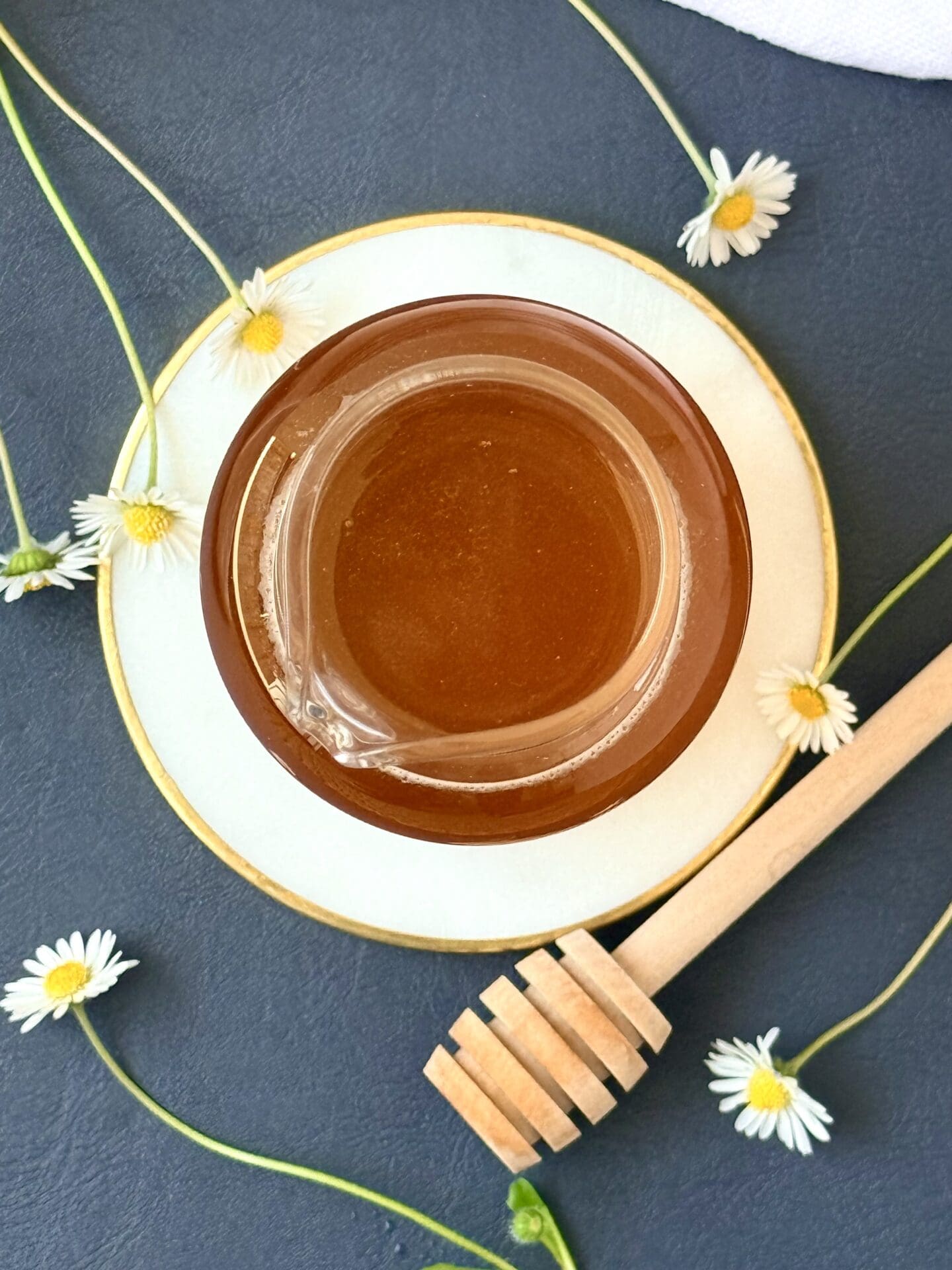 A glass bottle of honey simple syrup is seen from above, resting on a white and gold coaster.  Tiny white and yellow daisies and a wooden honey dipper surround the syrup.
