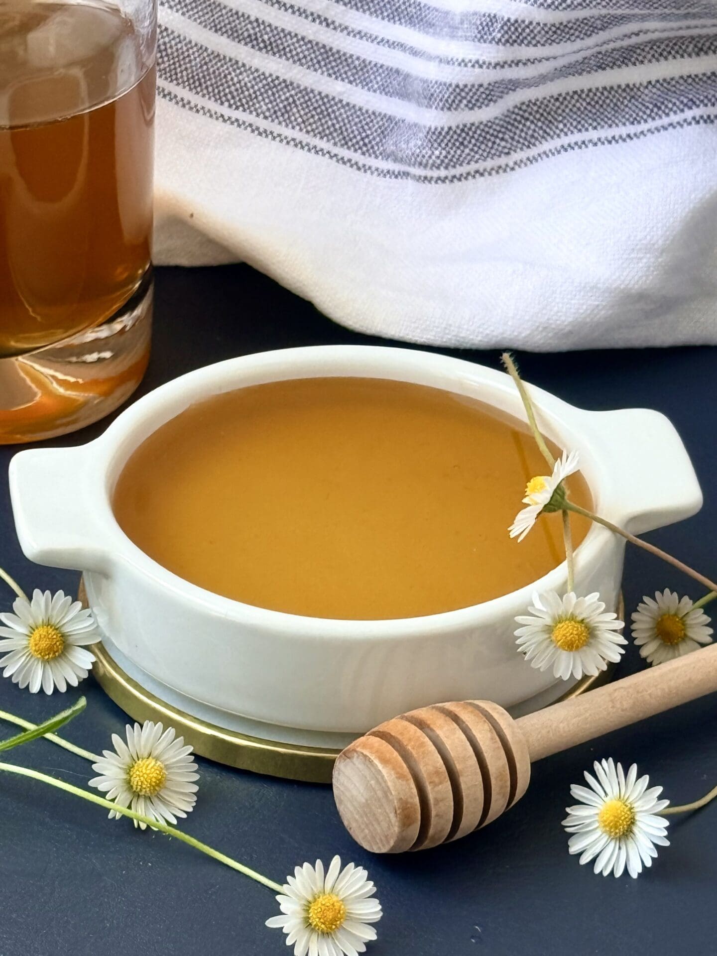A small white dish of honey simple syrup sits on a navy blue surface.  The bottle of syrup and a blue and white striped cloth sit in the background.  The dish is surrounded by small daisies.