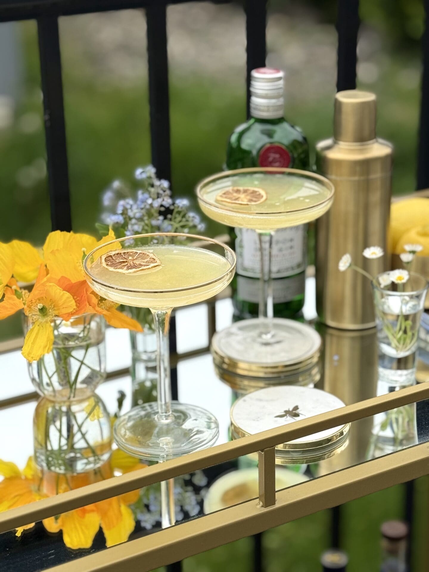 Two coupe glasses filled with Bee’s Knees cocktails sits on a gold and glass bar cart, set outdoors on a warm, sunny day.  The cocktails are surrounded by fresh flowers, a gold cocktail shaker and a bottle of gin.
