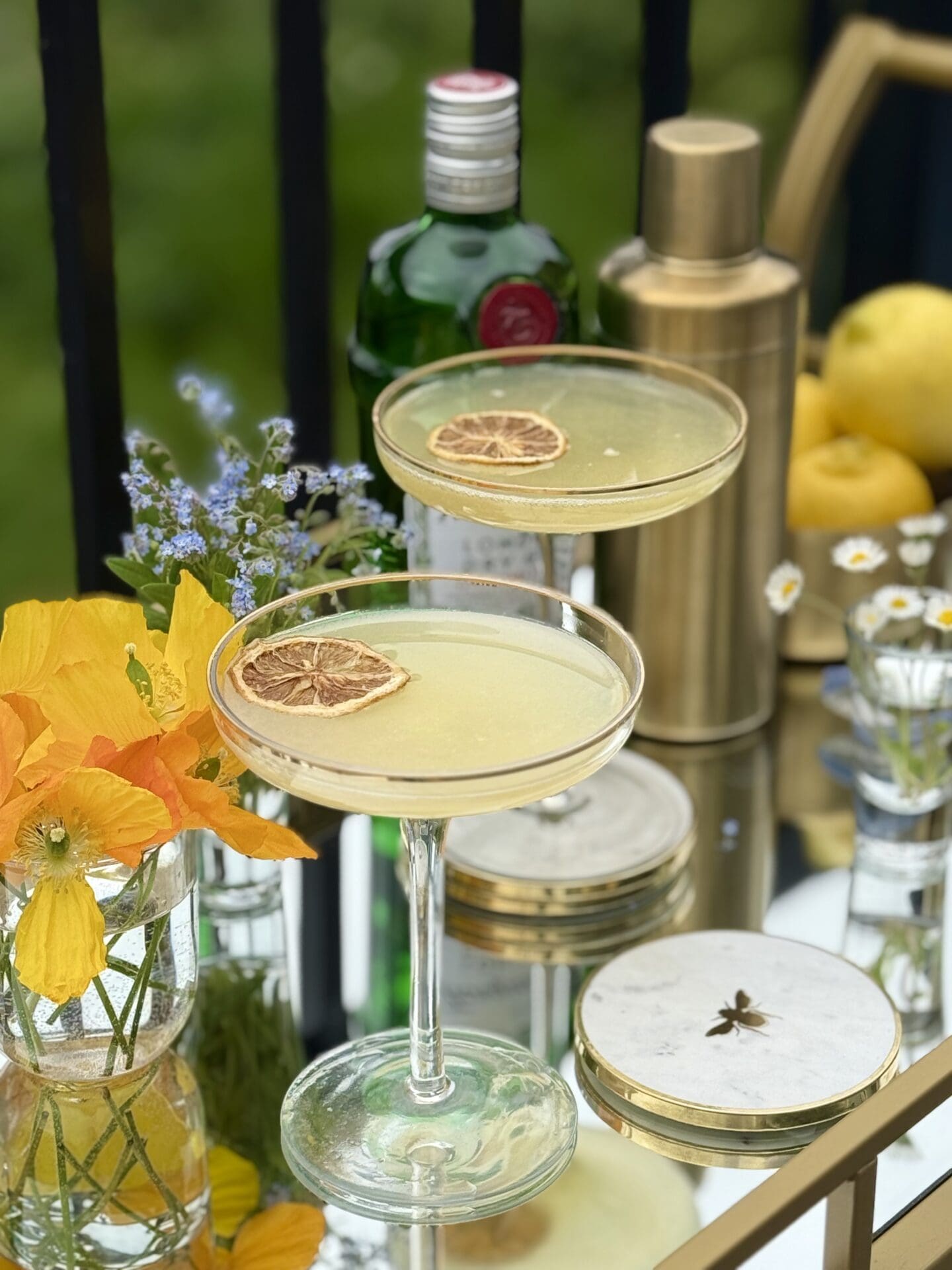 A bar cart filled with the ingredients to make a Bee’s Knees cocktail sits on a sunny patio.  Two coupe glasses filled with the drink are garnished with dried lemons slices and ready to be served.  