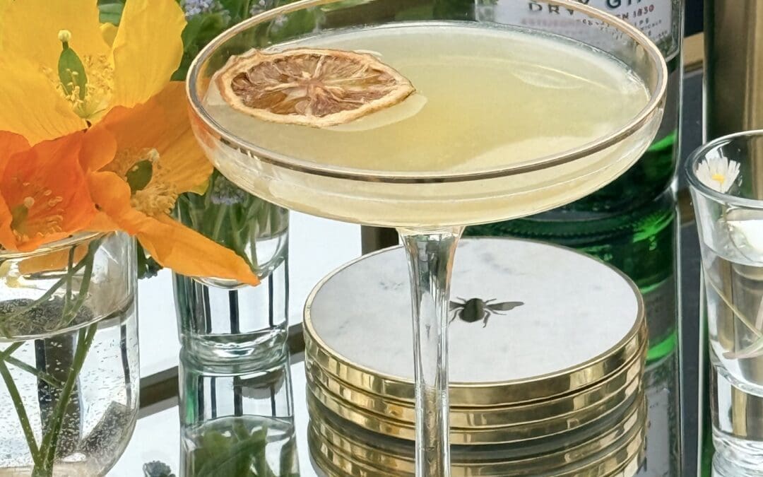 A coupe glass filled with a Bee’s Knees cocktail sits on a gold bar cart, surrounded by fresh flowers. A bottle of gin can be seen in the background.