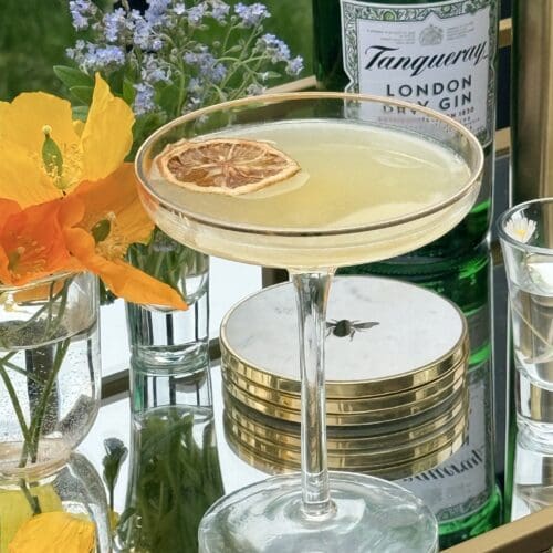 A coupe glass filled with a Bee’s Knees cocktail sits on a gold bar cart, surrounded by fresh flowers. A bottle of gin can be seen in the background.