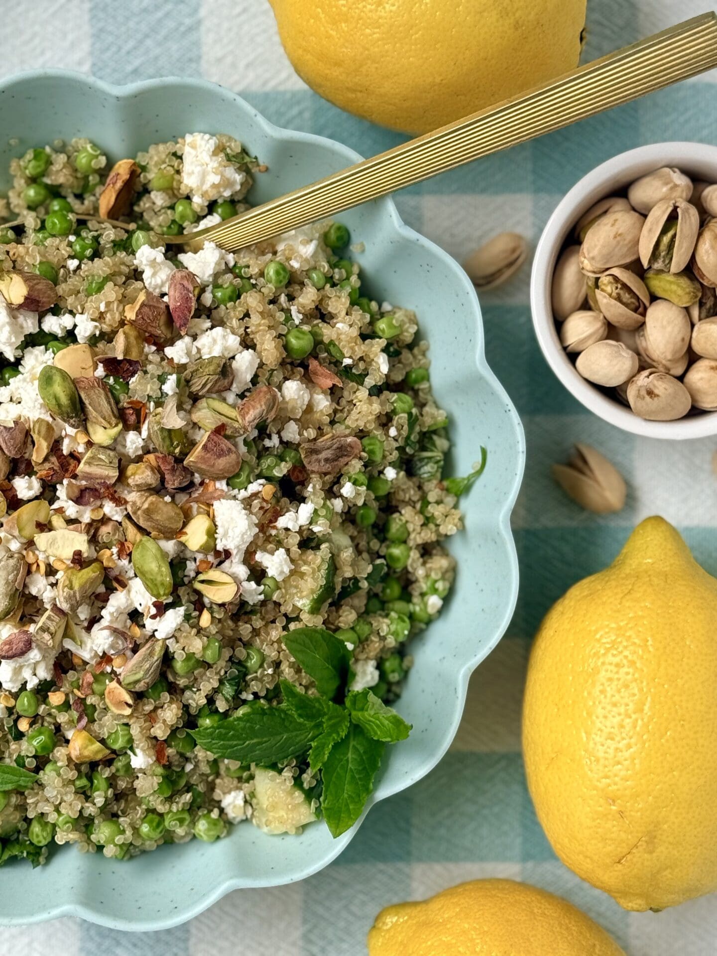 A turquoise bowl with a scalloped edge is filled with Lemony Quinoa Salad with Spring Peas and Pistachios.  The bowl rests on a checkered tablecloth and is surrounded by pistachios and lemons.