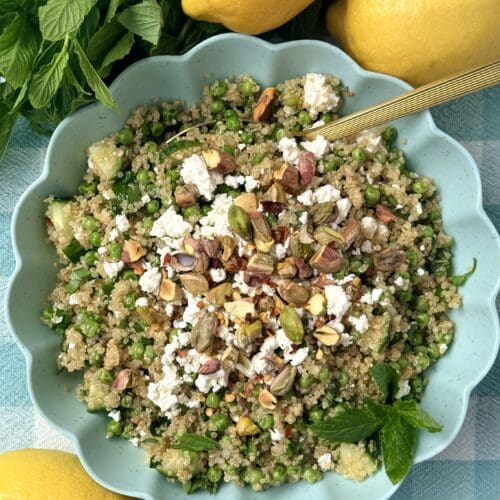 A bowl of Lemony Quinoa Salad with Spring Peas and Pistachios is seen from above, surrounded by fresh mint and lemons.