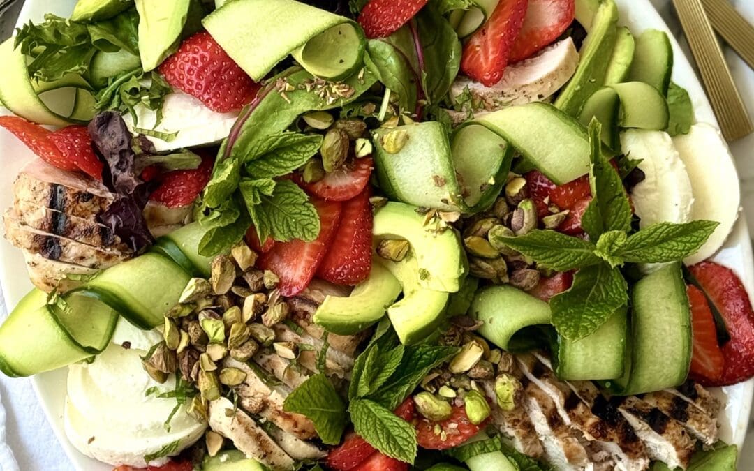 A large platter filled with Strawberry Cucumber Salad with Grilled Lemon Chicken is seen from above. The salad is beautifully arranged to show off its cucumber curls, bright red berries, creamy chunks of fresh mozzarella cheese and sprinkling of crunchy pistachios. A pair of gold-coloured salad servers sits nearby.