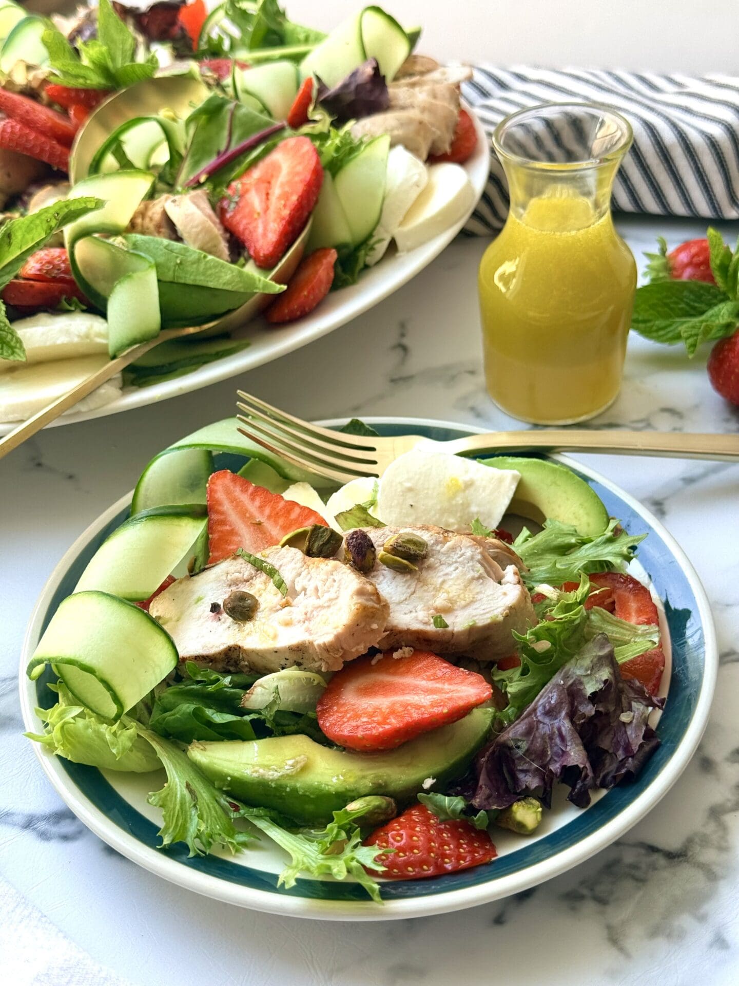 A plate of Strawberry Cucumber Salad with Grilled Lemon Chicken sits on a white marble counter, waiting to be eaten with a gold fork.  The platter of salad and bottle of lemon dressing are seen in the background.
