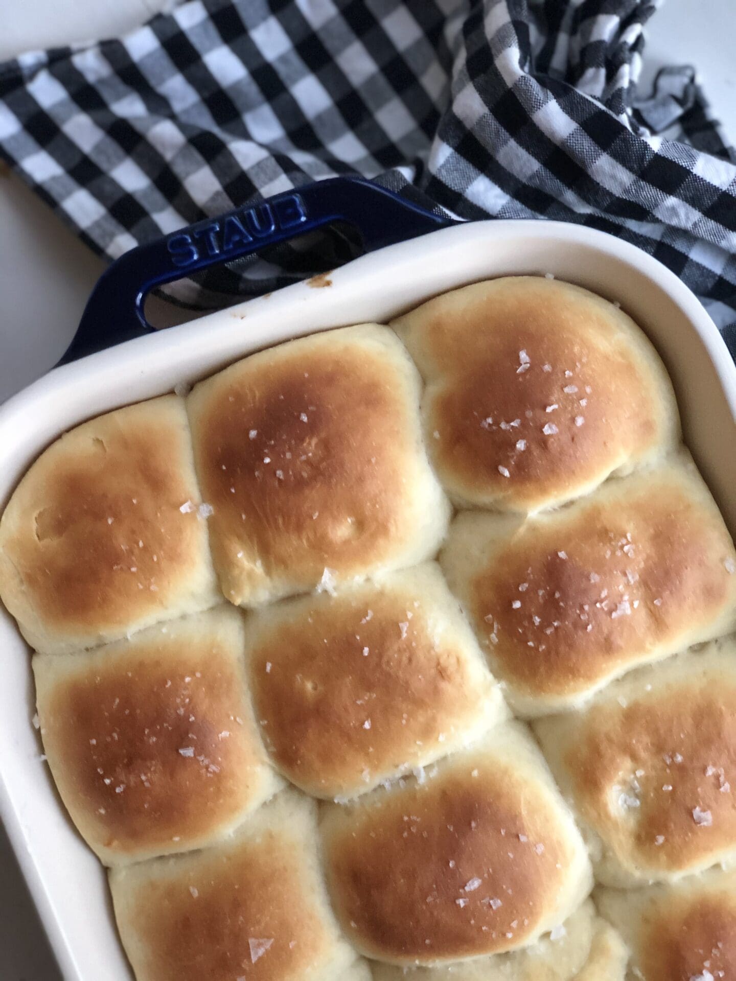 A pan of fresh-from-the-oven dinner rolls is seen from above, nestled together.  The tops of the buns are golden brown and sprinkled with flaky sea salt.