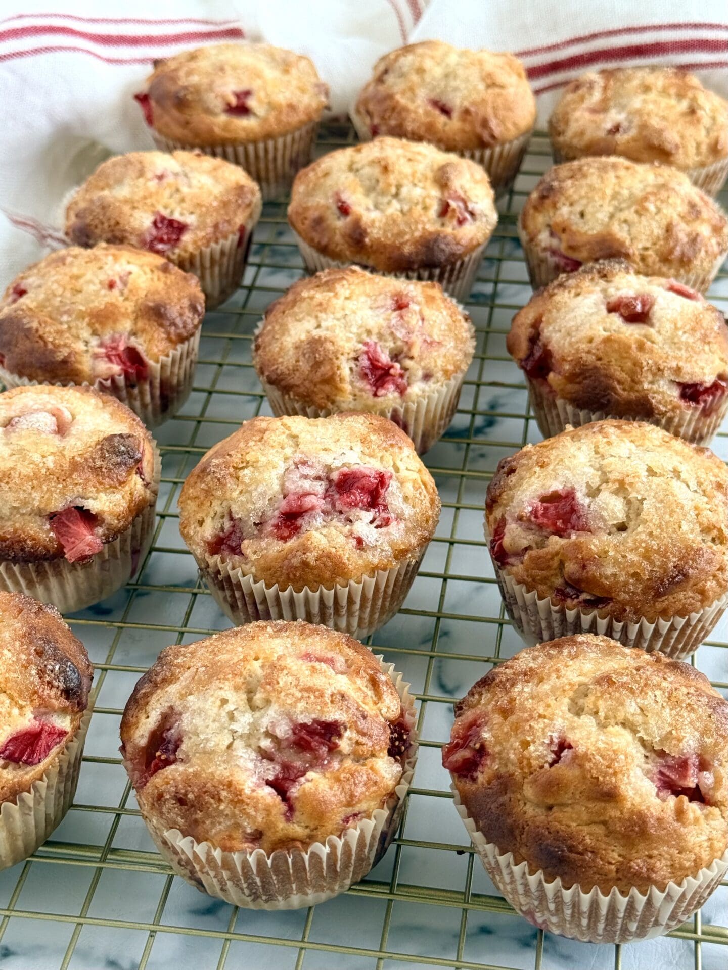 A batch of freshly baked strawberry yogurt muffins cools on a rack.  The muffins show off their crispy-sweet tops studded with fresh strawberries.