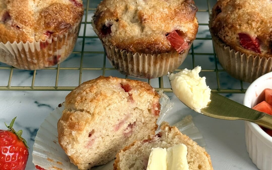 A strawberry yogurt muffin sits in the foreground, split in half and slathered in butter. The rest of the batch of muffins cools on a rack in the background. Juicy red strawberries decorate the scene.