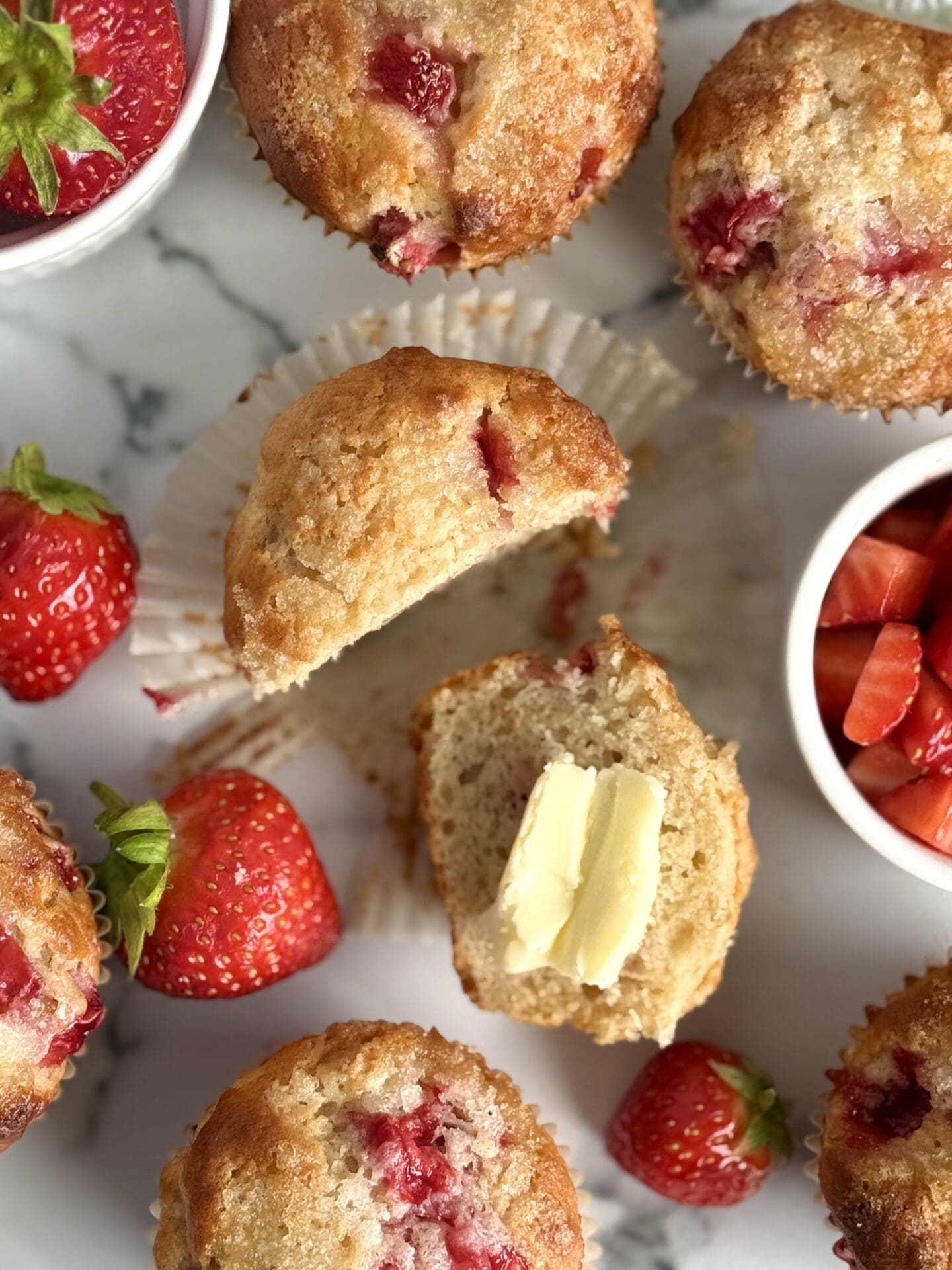 A strawberry yogurt muffin is seen from above, split in two and slathered in butter.  Fresh strawberries and more golden-topped muffins complete the scene.