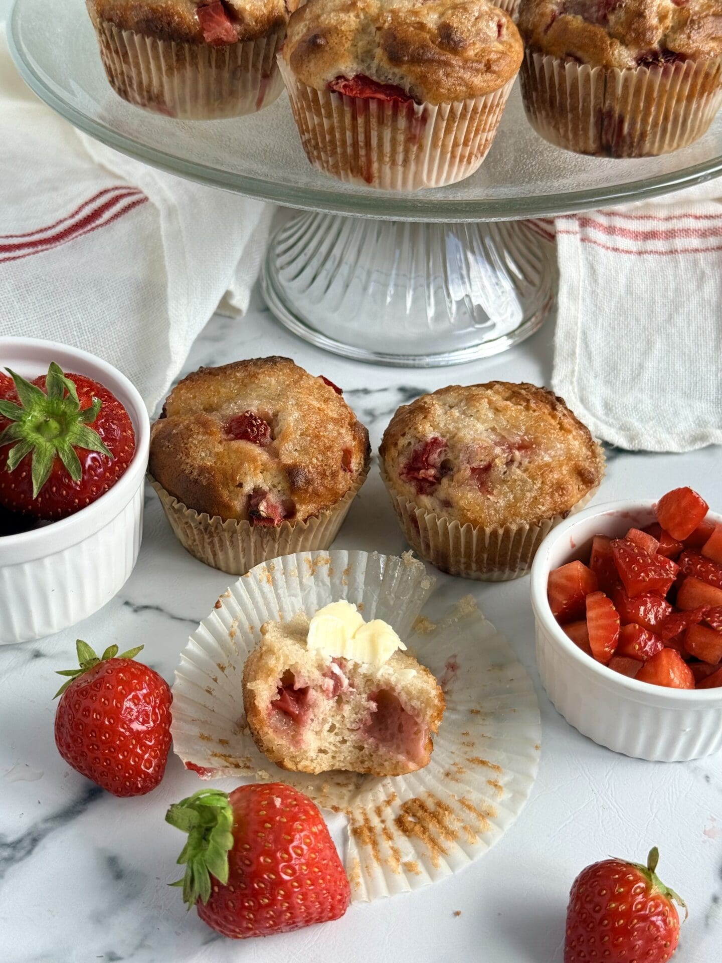A large bite has been taken out of a freshly baked, butter-topped,  strawberry yogurt muffin, revealing its strawberry studded interior.  A glass cake plate filled with muffins and bowls of fresh strawberries are seen in the background.