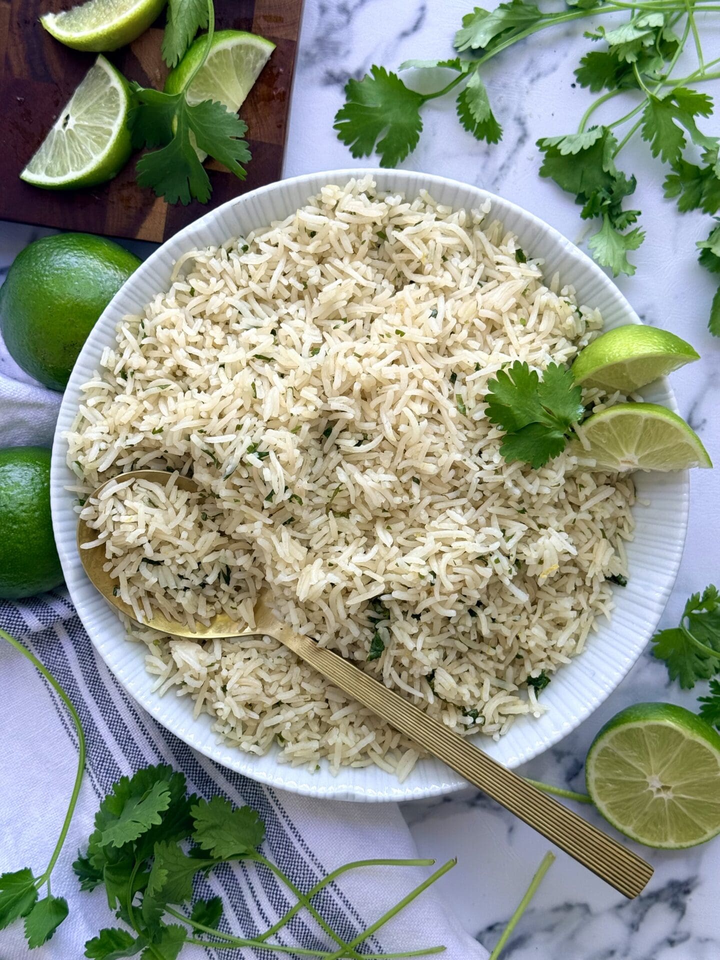 A large bowl of cilantro lime rice sits in the centre of the frame.  The bowl rests on a white marble counter top.  A gold coloured serving spoon is seen scooping a large serving.  The rice is surrounded by fresh lime slices and cilantro leaves.