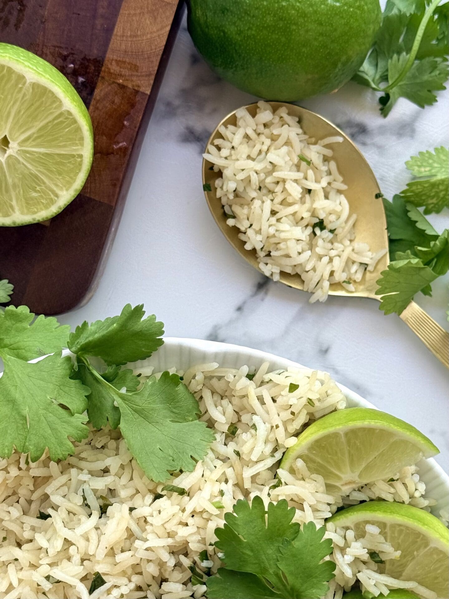 A gold coloured serving spoon is filled with a scoop of cilantro lime rice.  The spoon rests on a white marble counter near a dark wood cutting board.  Fresh lime and cilantro complete the scene.
