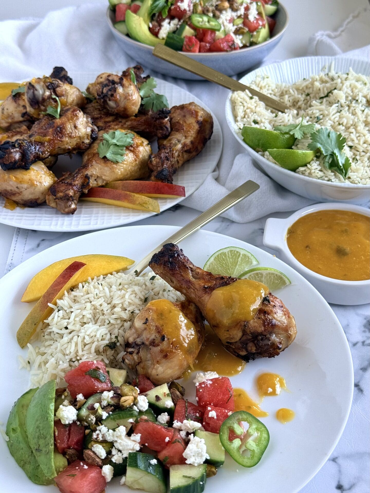 A dinner plate is seen in the foreground, filled with a serving of vibrant watermelon avocado salad, mango glazed chicken drumsticks and cilantro lime rice.  Full platter of each of these dishes is seen in the background.