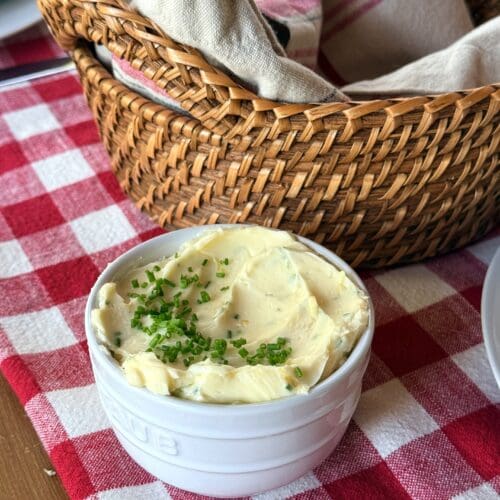 A small bowl of Lemon Chive Butter sits on a red and white gingham tablecloth, near a basket of fresh bread. The top of the butter is generously garnished with chopped fresh chives.