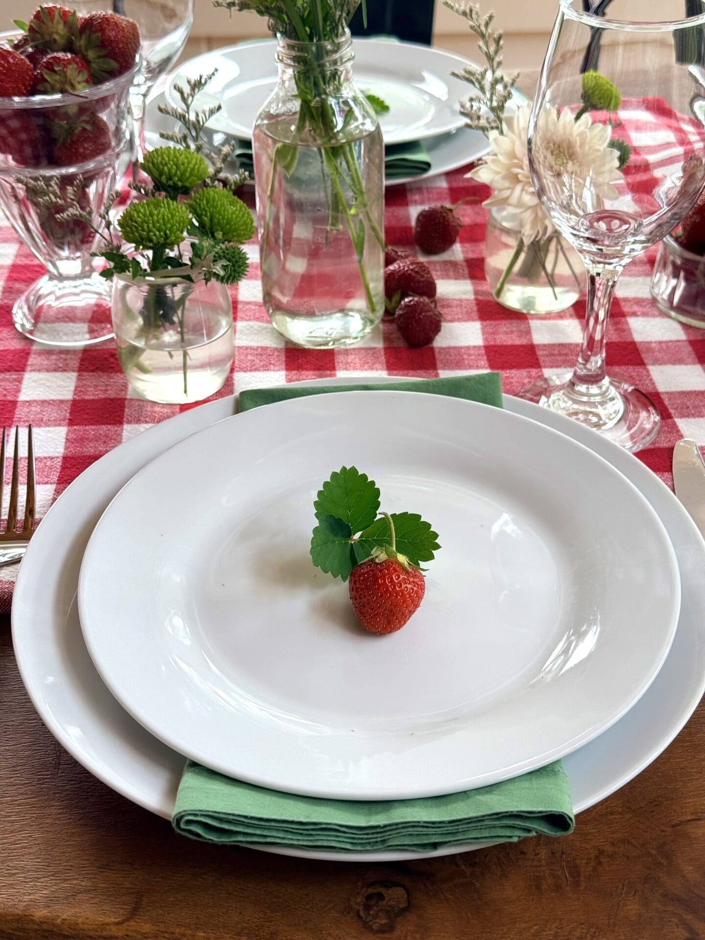 An individual place setting at a Strawberry Social lunch is the focus of this photo.  The place setting features white plates, a green cloth napkin, stemware and cutlery.  The plate is finished with a plump strawberry with strawberry leaves.  The place setting rests  on a red gingham tablecloth.  Small vases of white and green wild flowers are seen in the background.