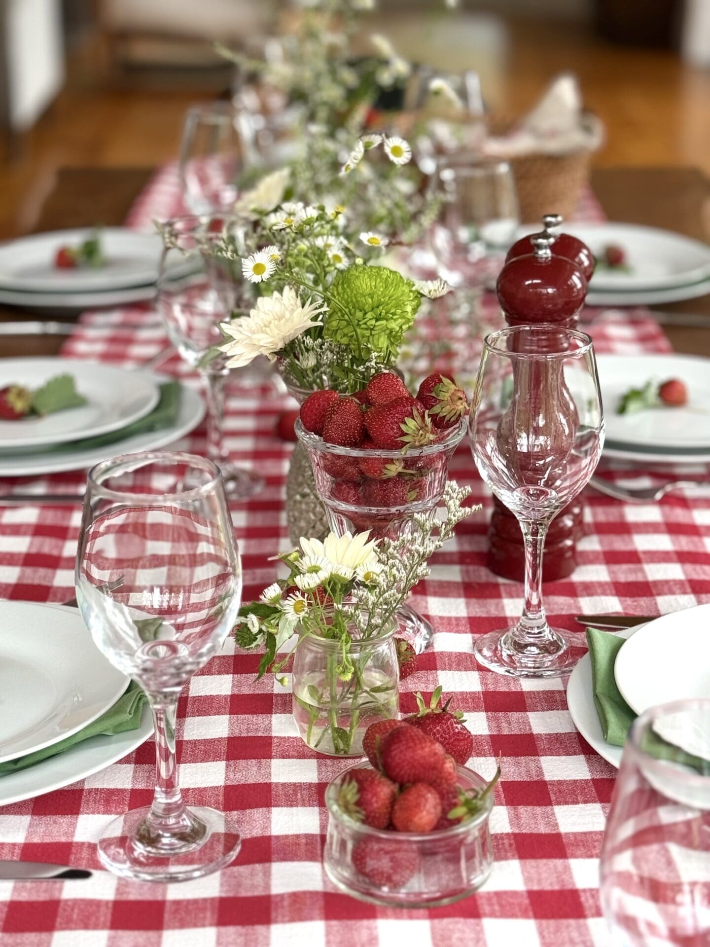 A table is set with a red gingham tablecloth and vases of wild flowers for a Strawberry Social lunch. Bowls of fresh strawberries spill out onto the table.