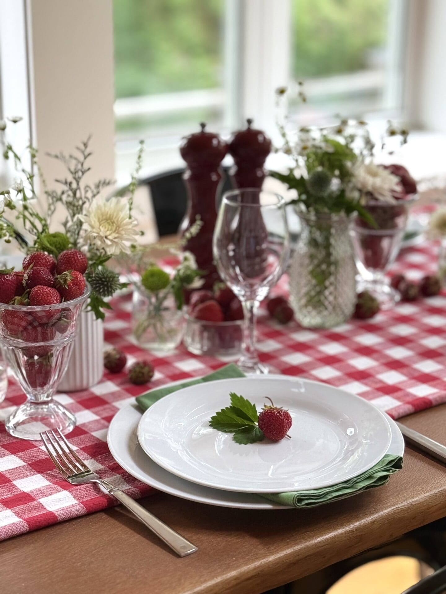 The table is set in a sun-filled room for a Strawberry Social lunch.  The table features a red and white gingham table cloth, mismatched vases of white and green wild flowers and plenty of fresh strawberries.