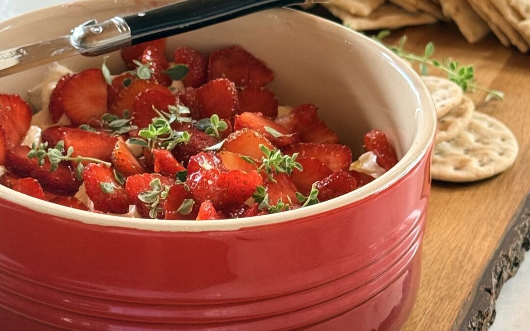 A red bowl filled with Whipped Brie with Strawberries and Fresh Thyme sits on a wood board surrounded by crackers.