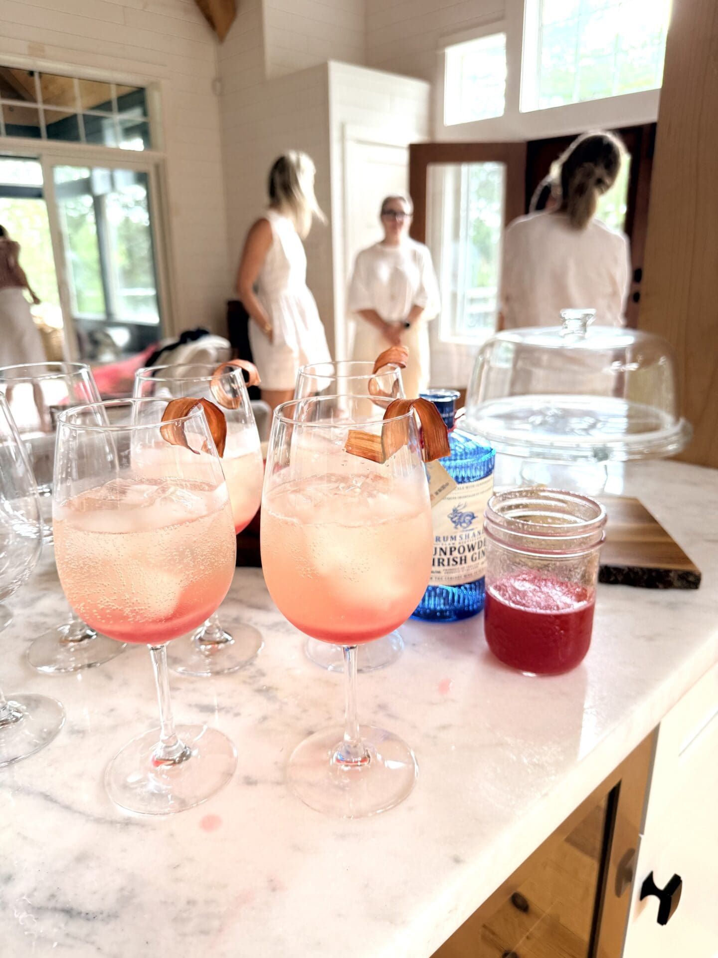 A group of Rhubarb Gin Spritz Cocktails sit on a marble bar.  Party guests are seen in conversation in the background.