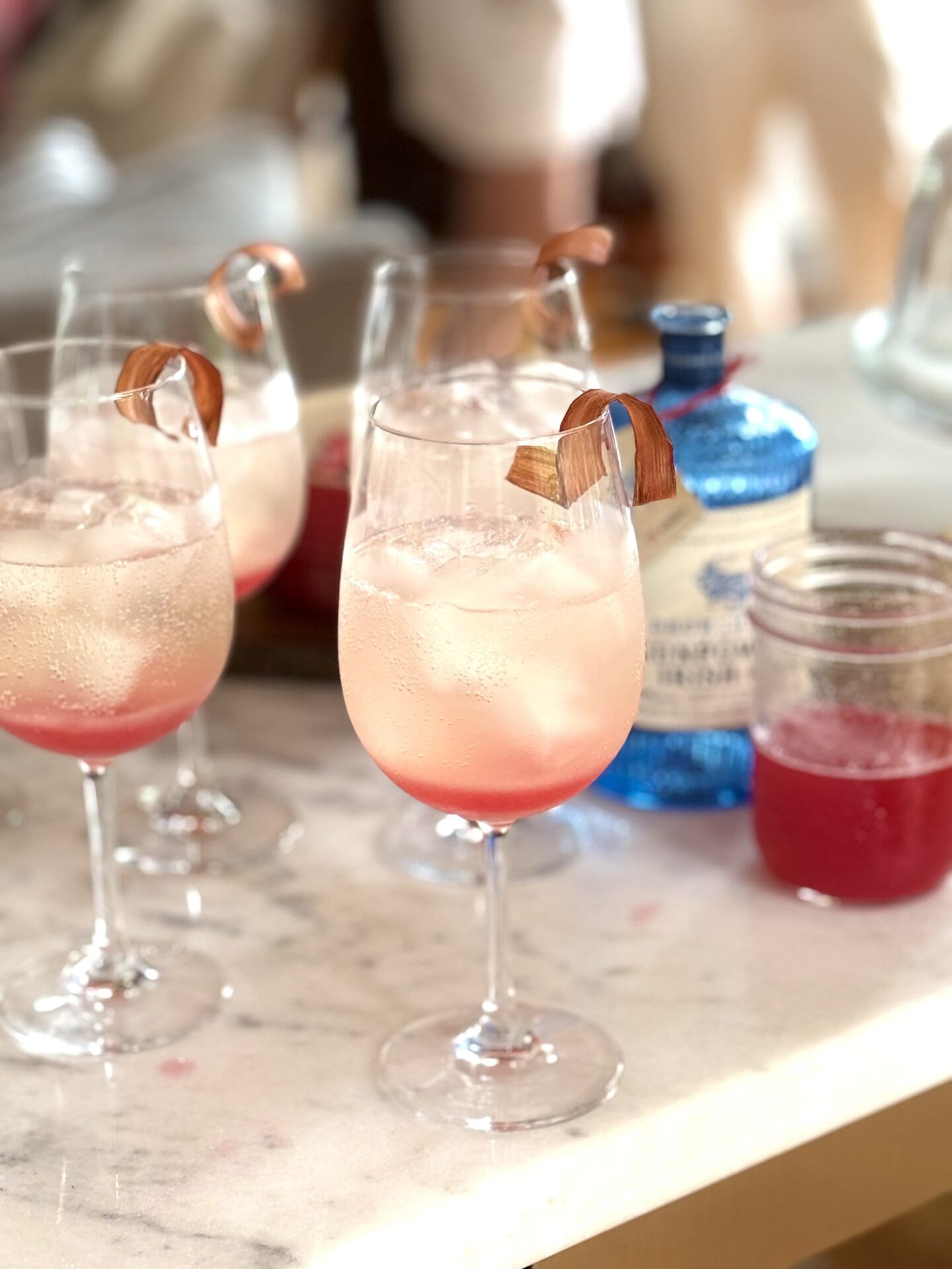 Four Rhubarb Gin Spritz cocktails are being made on a white marble bar.  A blue bottle of gin is seen in the background, along with a jar of ruby red rhubarb syrup.