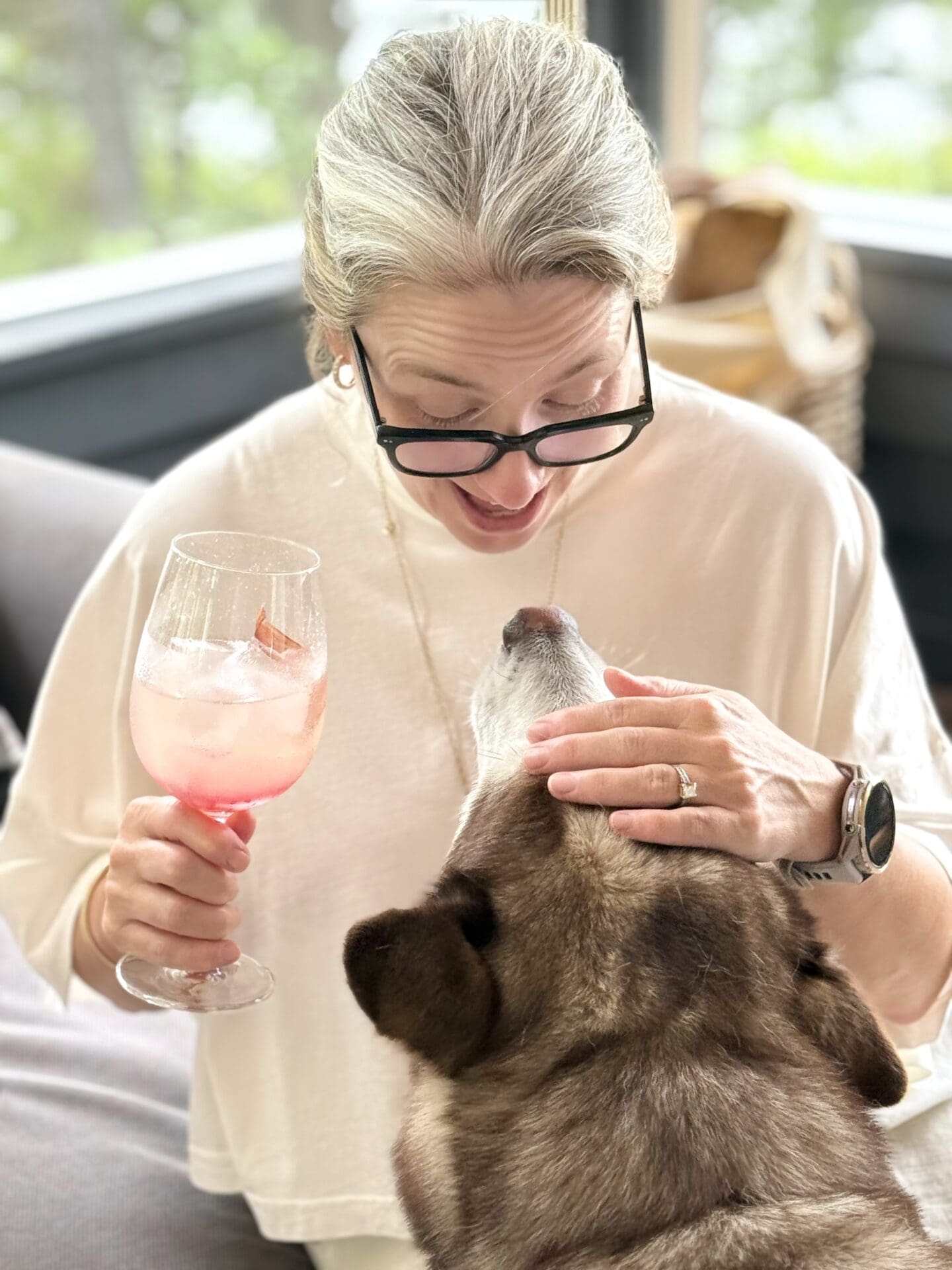 A woman looks down at and lovingly pets a husky while the dog looks back up at her.  She is holding a cocktail and they are sitting on a breezy screened in porch.