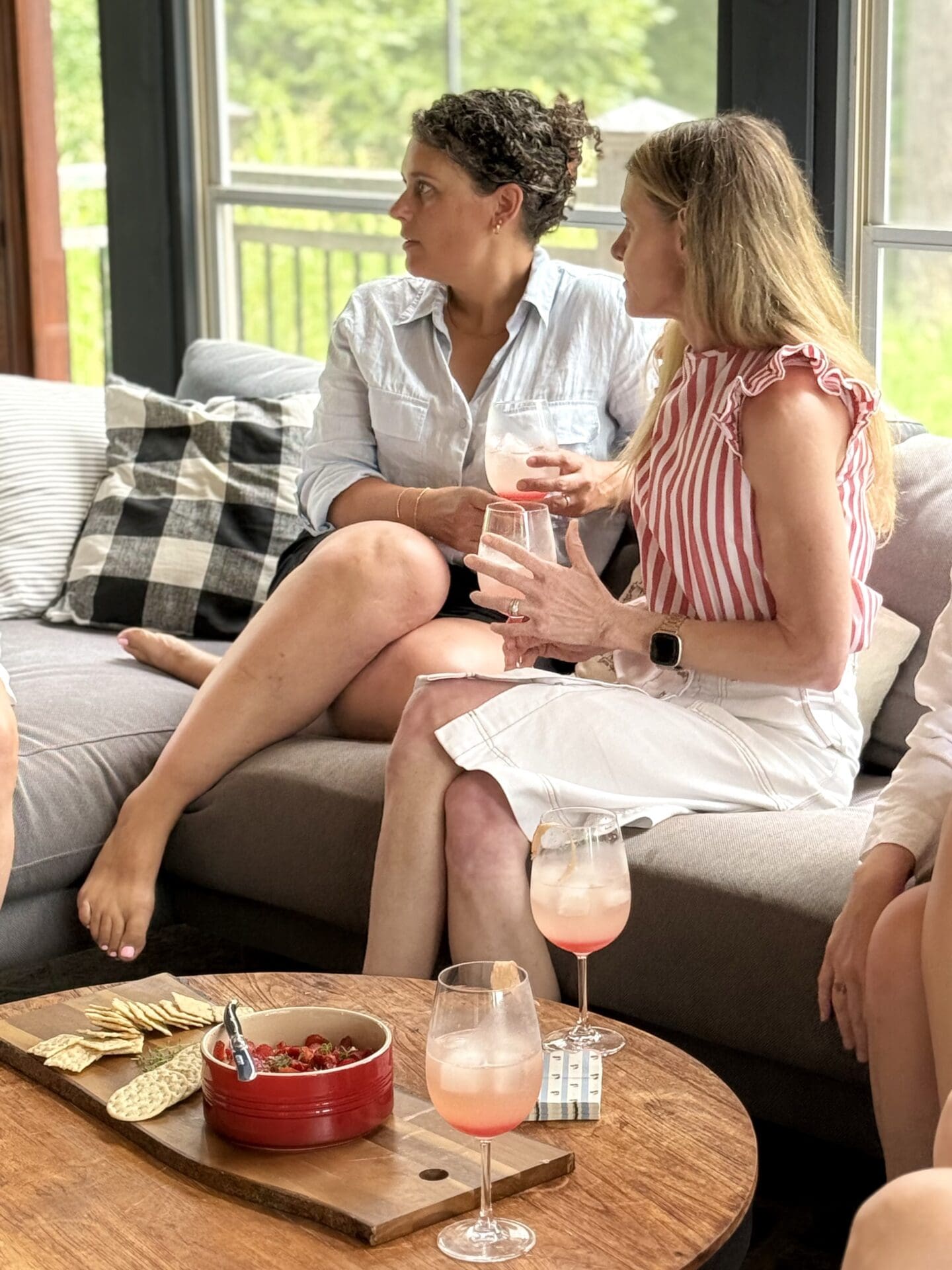 Two women sit on a screened in porch, deep in conversation while enjoying a cocktail and appetizer.