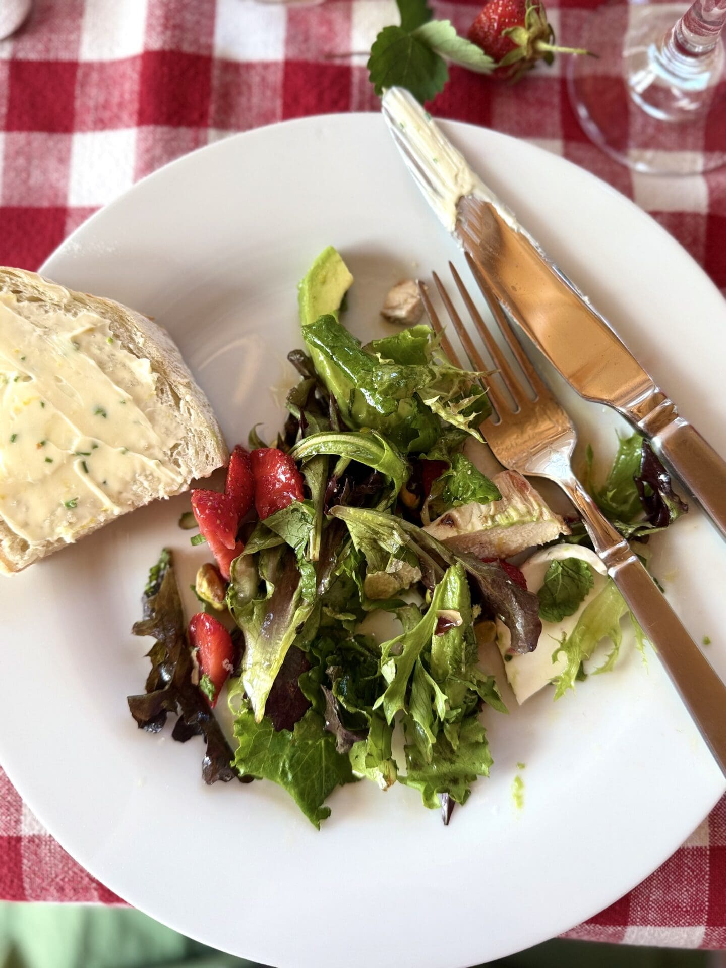 A dinner plate containing salad is seen from above.  A slice of bread, slathered in lemon chive butter perches on the edge of the plate.  The plate rests on a red gingham tablecloth.