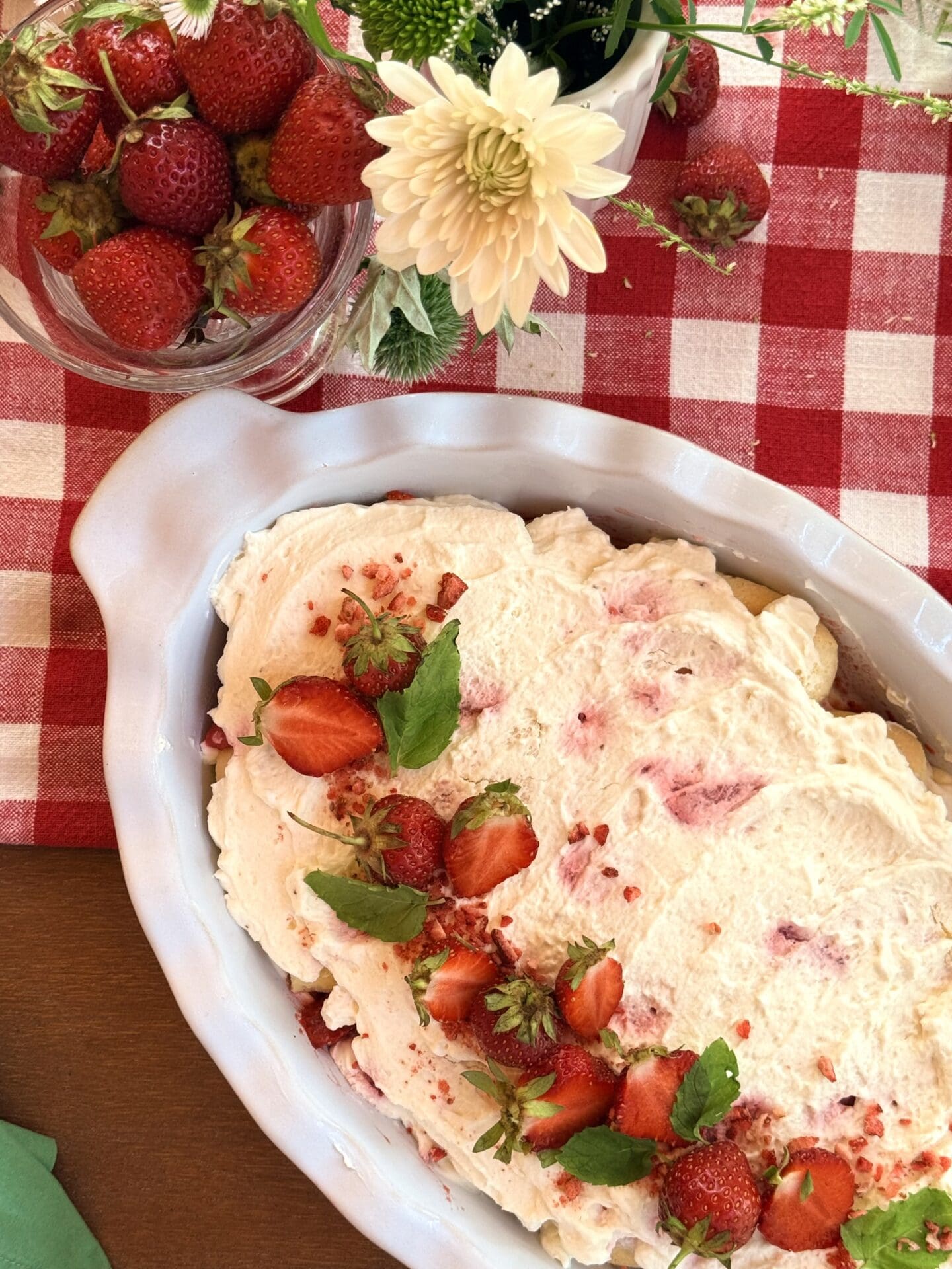 A large serving dish of strawverry tiramisu is seen from above, resting on a red and white tablecloth.  The tiramisu is beautifully garnished with fresh strawberries and mint.