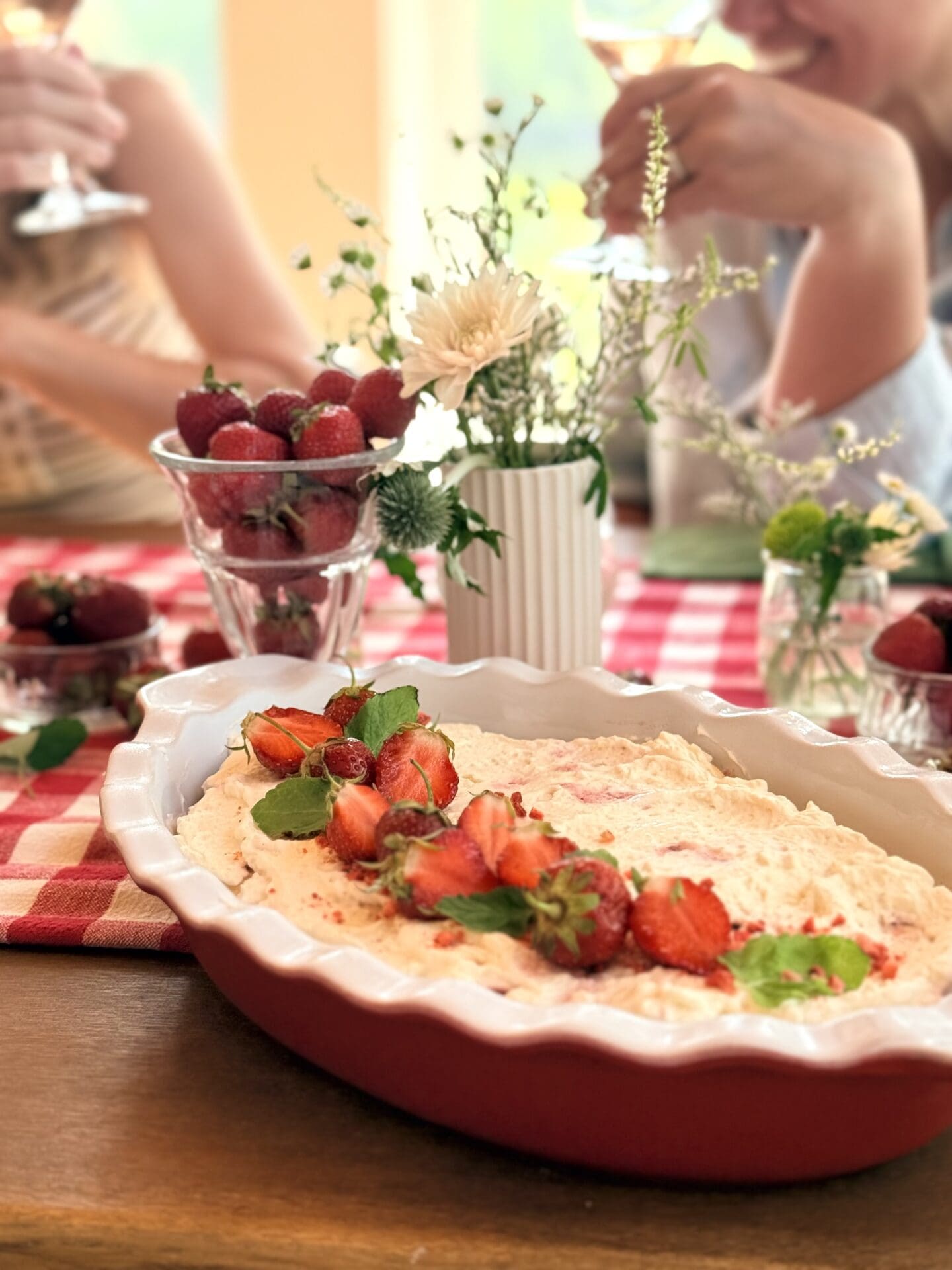 A dish of Strawberry Tiramisu sits on a table set for lunch. Guests are seen in conversation in the background.