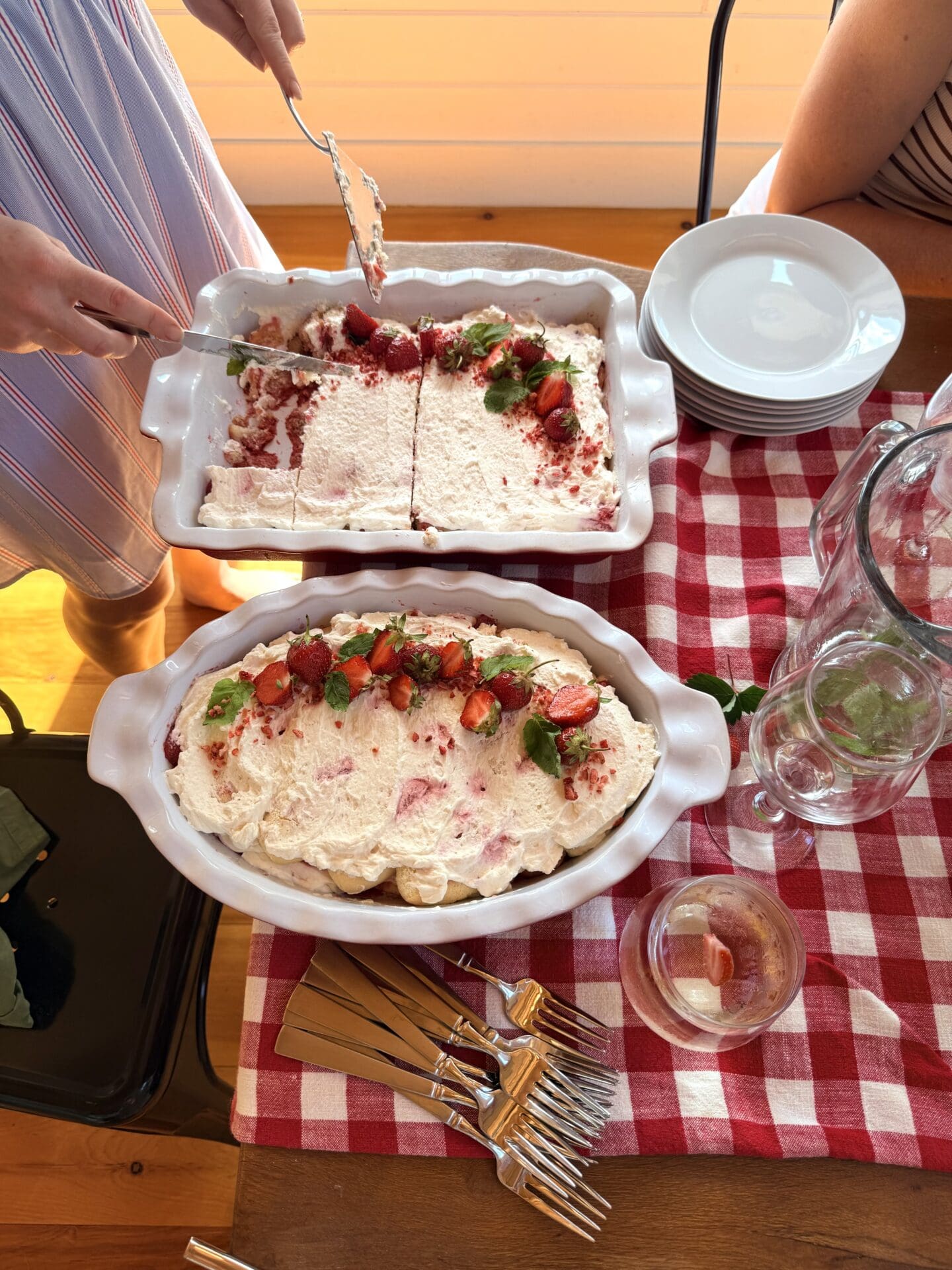 A woman in a blue dress serves strawberry tiramisu from two large dishes at a strawberry social lunch.