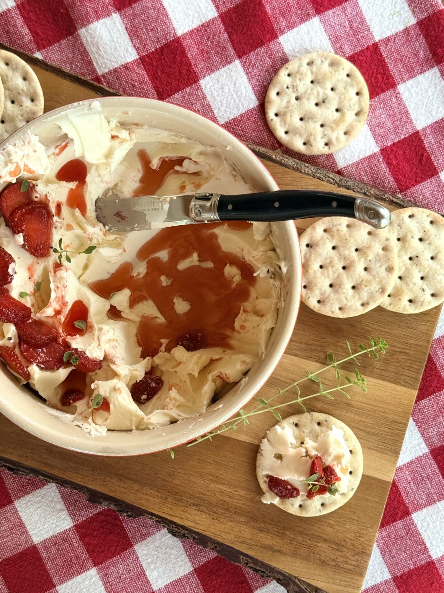 A bowl of whipped Brie with stawberries and thyme is seen from above on a red gingham tablecloth.  The appetizer is being devoured, showing the swoops of whipped cheese under the layer of fresh strawberries.  A cracker with a generous amount of the whipped Brie sits beside the bowl, ready to be eaten.