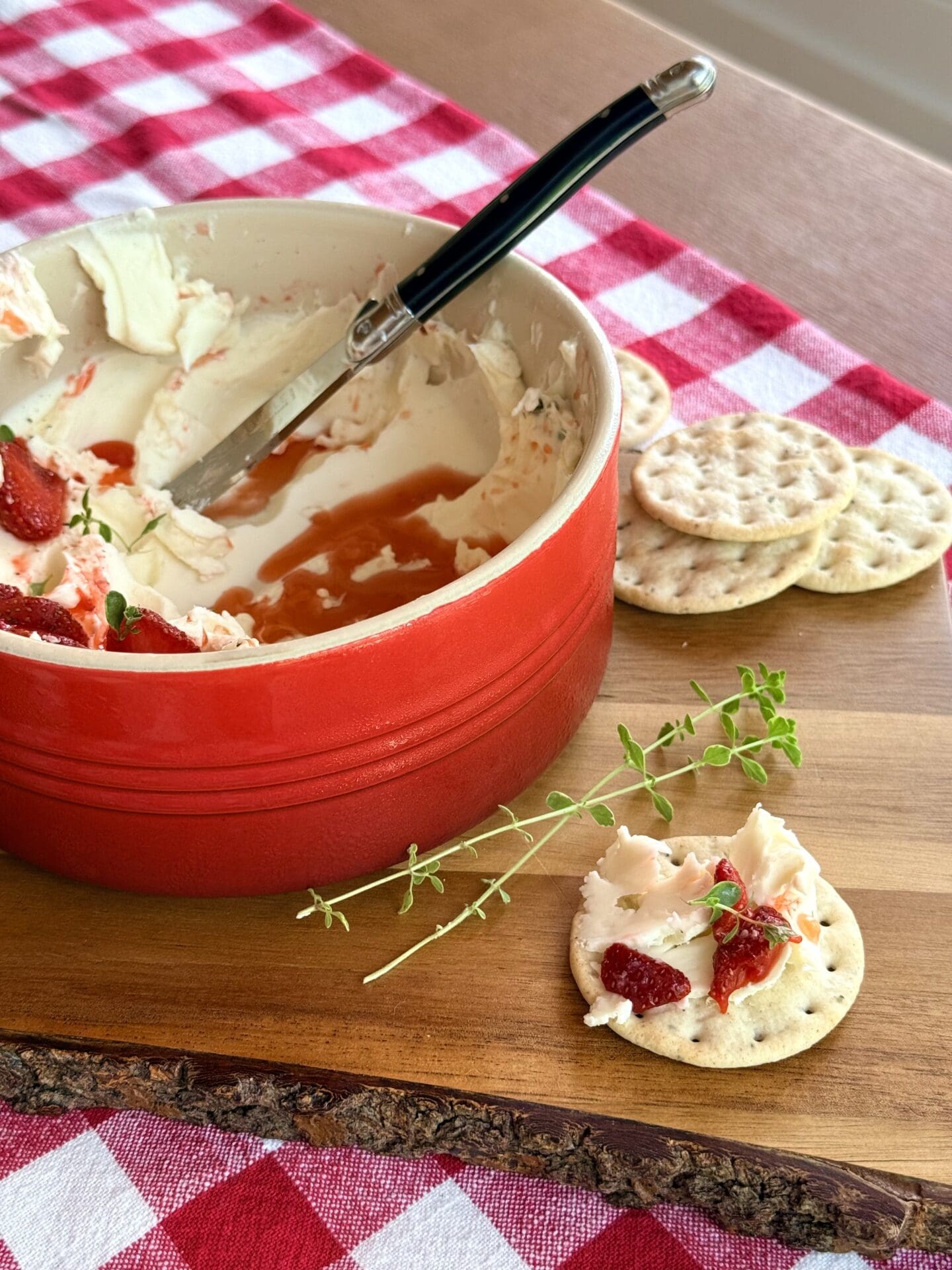 A bowl of Whipped Brie with Strawberries and Fresh Thyme sits on a wood board.  The appetizer has been partially enjoyed, showing the swoops of creamy cheese and juicy strawberry topping.  A cracker sits in the foreground, topped with the brie and strawberry mixture.