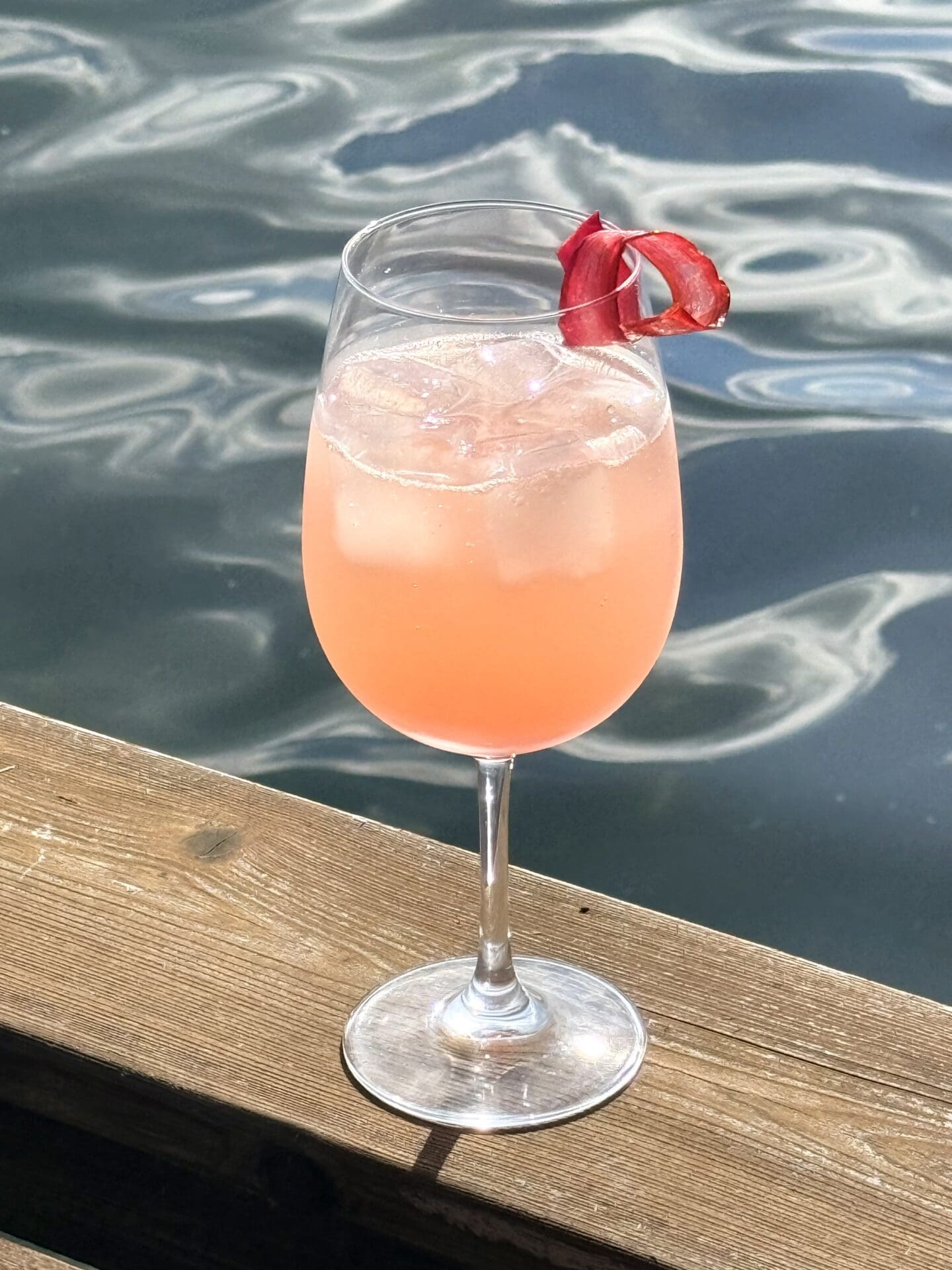 A large stemmed glass is filled with an ice-cold,  blush pink, Rhubarb Gin Spritz cocktail.  The drink rests on a wood railing, overlooking a sparkling lake.