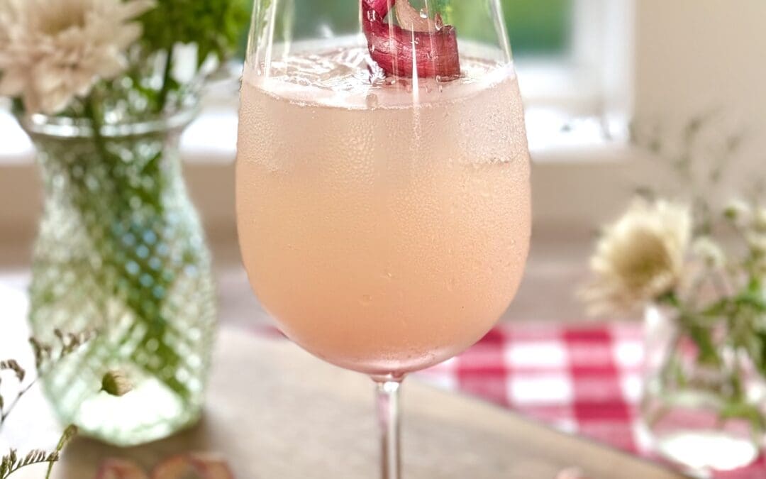 A rhubarb gin spritz cocktail sits on a wood board in front of a window. A summery scene of trees and a lake can be seen through the window. The drink is surrounded by bouquets of wildflowers. A red gingham tablecloth is seen in the background.