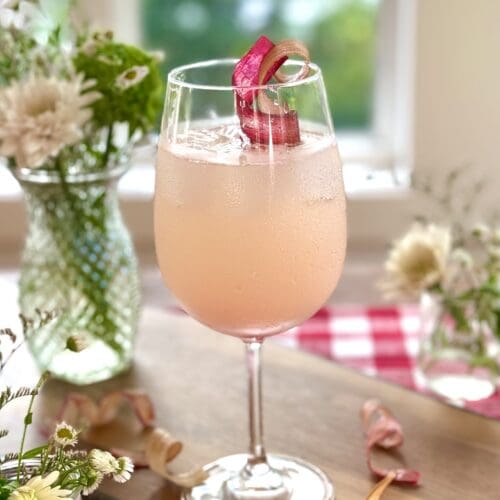 A rhubarb gin spritz cocktail sits on a wood board in front of a window. A summery scene of trees and a lake can be seen through the window. The drink is surrounded by bouquets of wildflowers. A red gingham tablecloth is seen in the background.