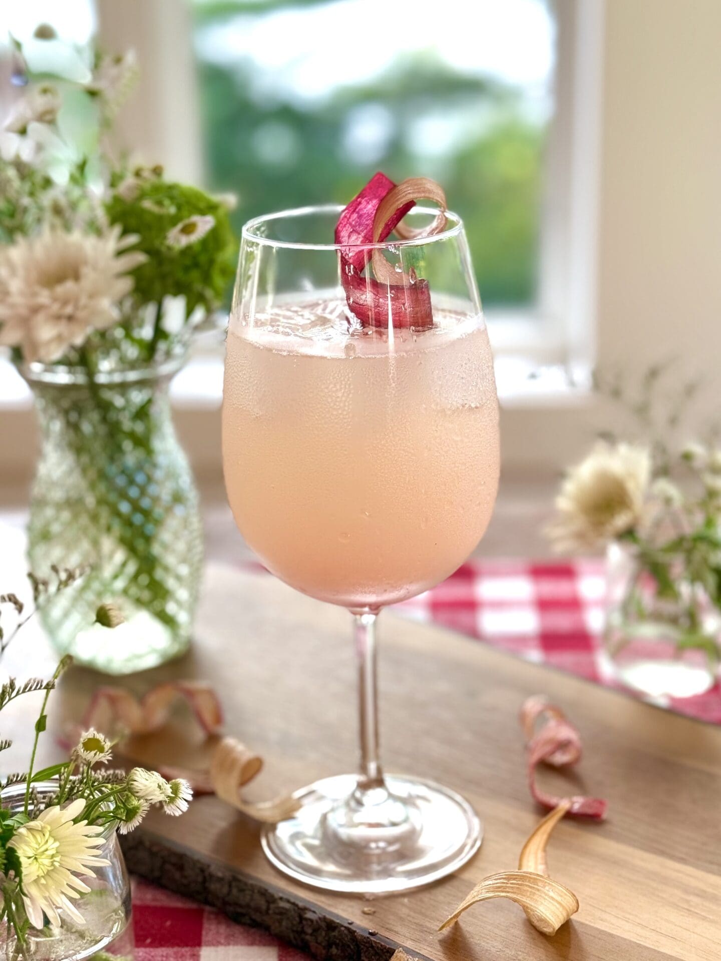 IMG_9608 A rhubarb gin spritz cocktail sits on a wood board in front of a window. A summery scene of trees and a lake can be seen through the window. The drink is surrounded by bouquets of wildflowers. A red gingham tablecloth is seen in the background.
