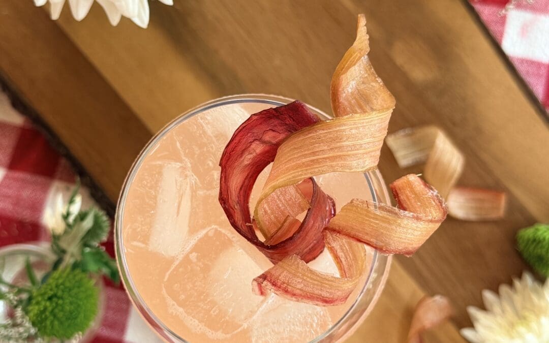 A cocktail glass is seen from above, filled with an ice cold pink drink and garnished lavishly with candied rhubarb ribbons. The glass sits on a wood board, surrounded by flowers. A red gingham tablecloth completes the image.