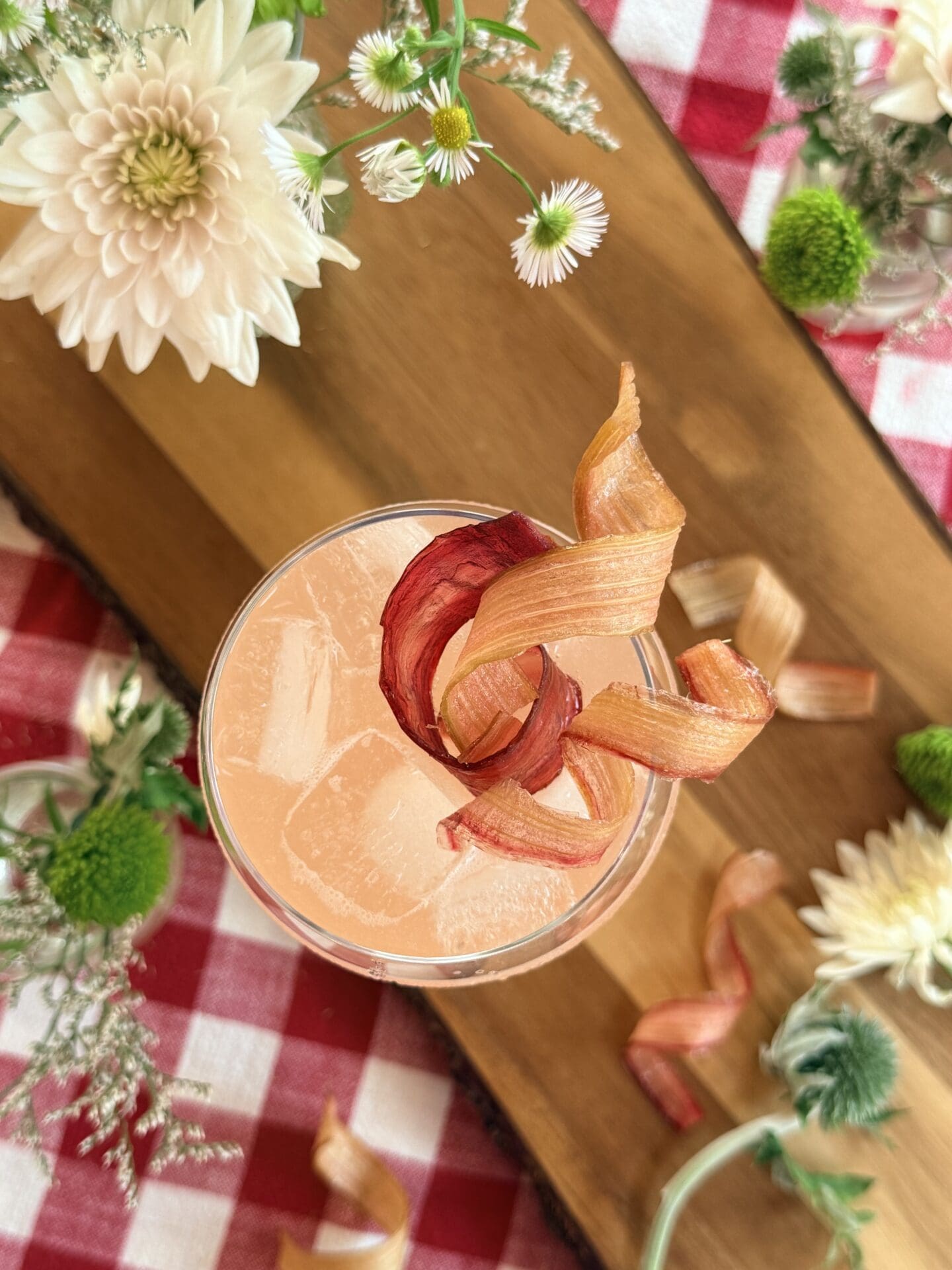 IMG_9618 A cocktail glass is seen from above, filled with an ice cold pink drink and garnished lavishly with candied rhubarb ribbons. The glass sits on a wood board, surrounded by flowers. A red gingham tablecloth completes the image.