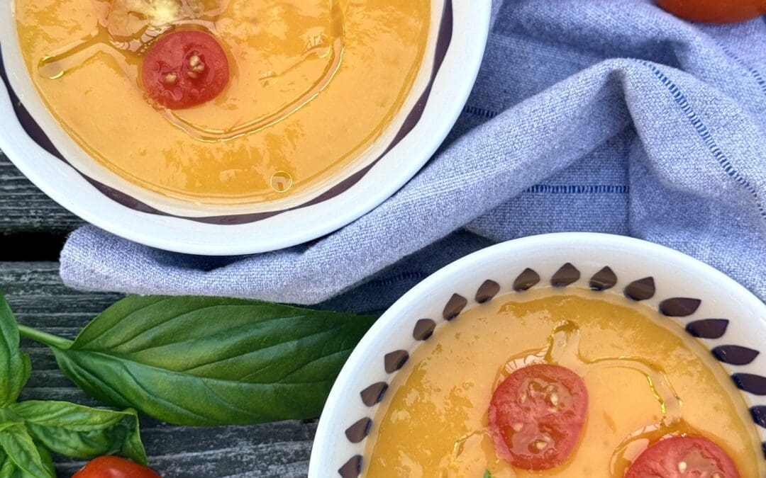 Two bowls of Yellow Tomato Gazpacho are seen from above, showcasing its beautiful sunny yellow colour. The bowls of soup are garnished with sliced cherry tomatoes and basil leaves.