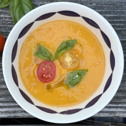 A single bowl of bright Yellow Tomato Gazpacho is seen from above on a wood table. The soup is garnished with cherry tomatoes and fresh basil.